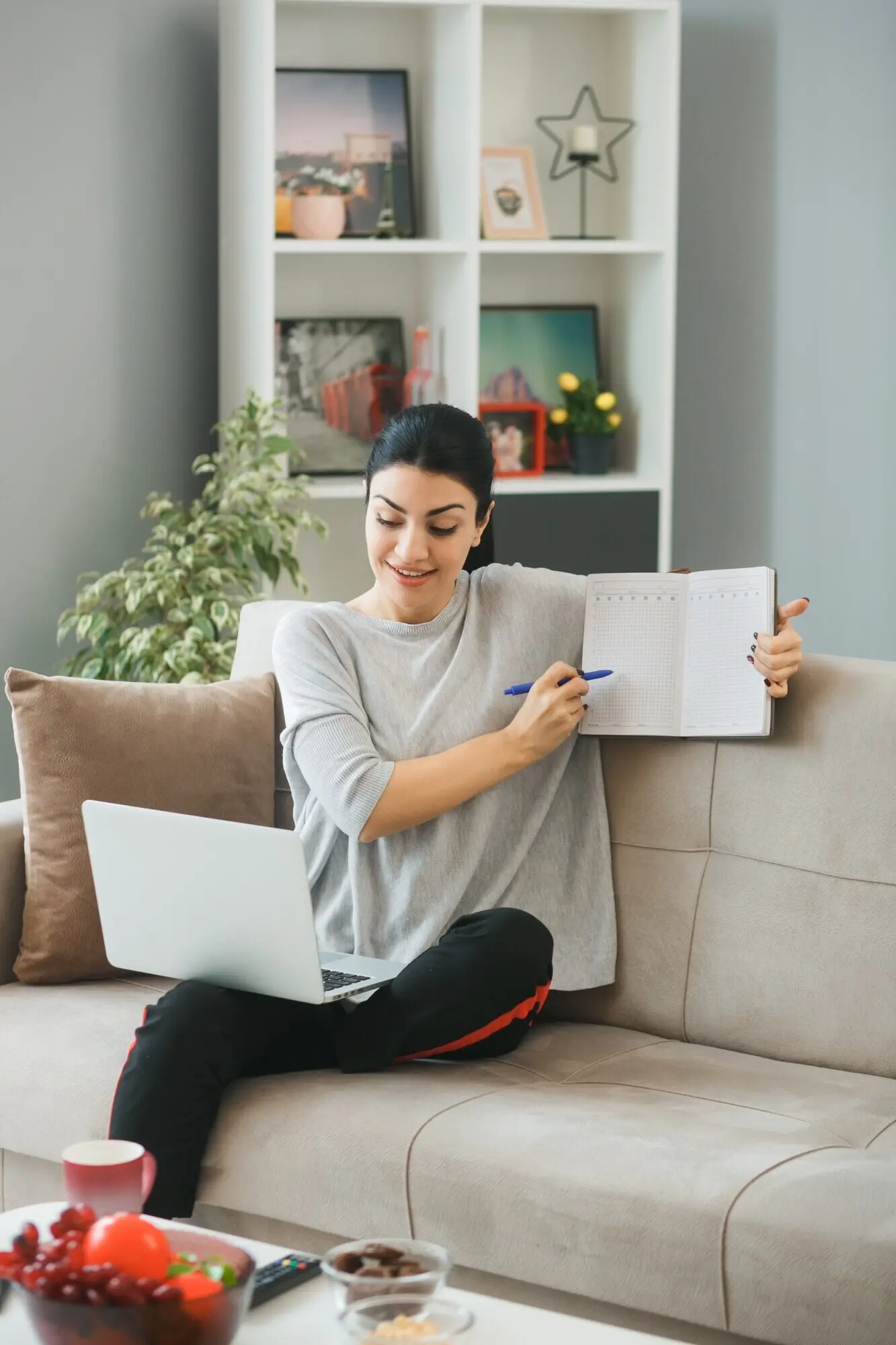 A smiling young girl sits on a sofa in a living room, using a laptop and holding a pen to point at a book.